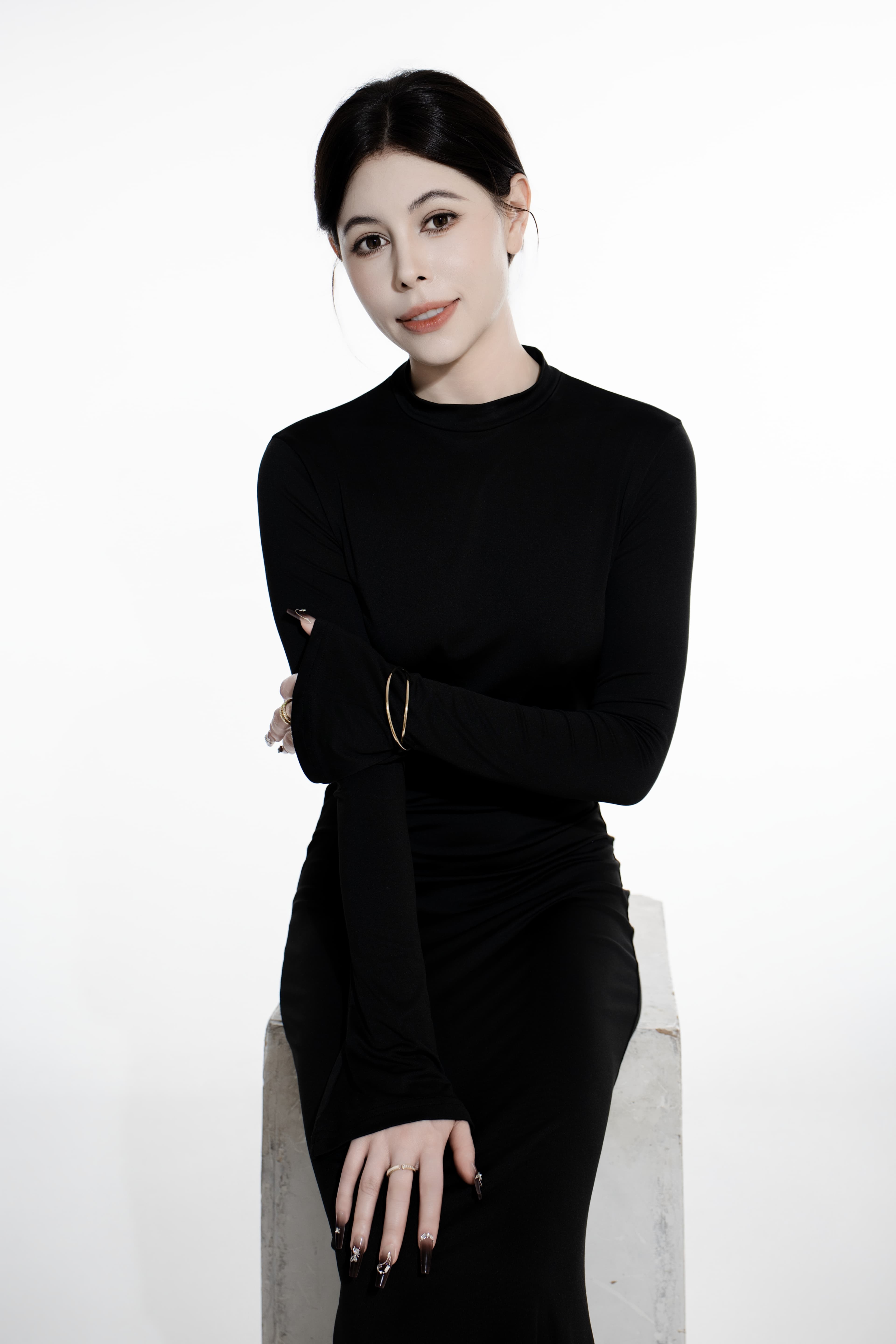 Woman in a black long-sleeved dress sitting on a white block against a white background.