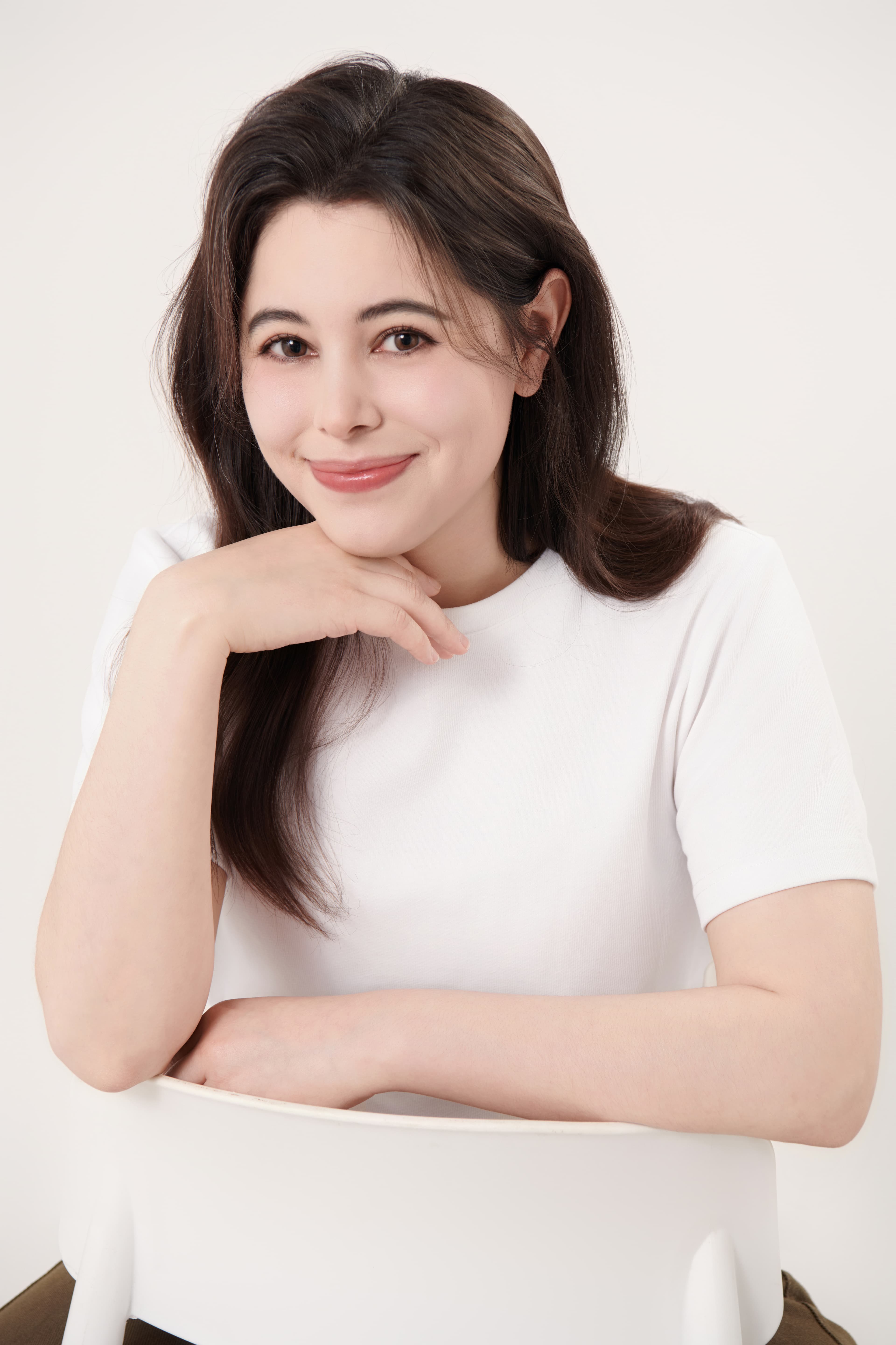 Smiling woman with long dark hair wearing a white t-shirt, resting chin on her hand.